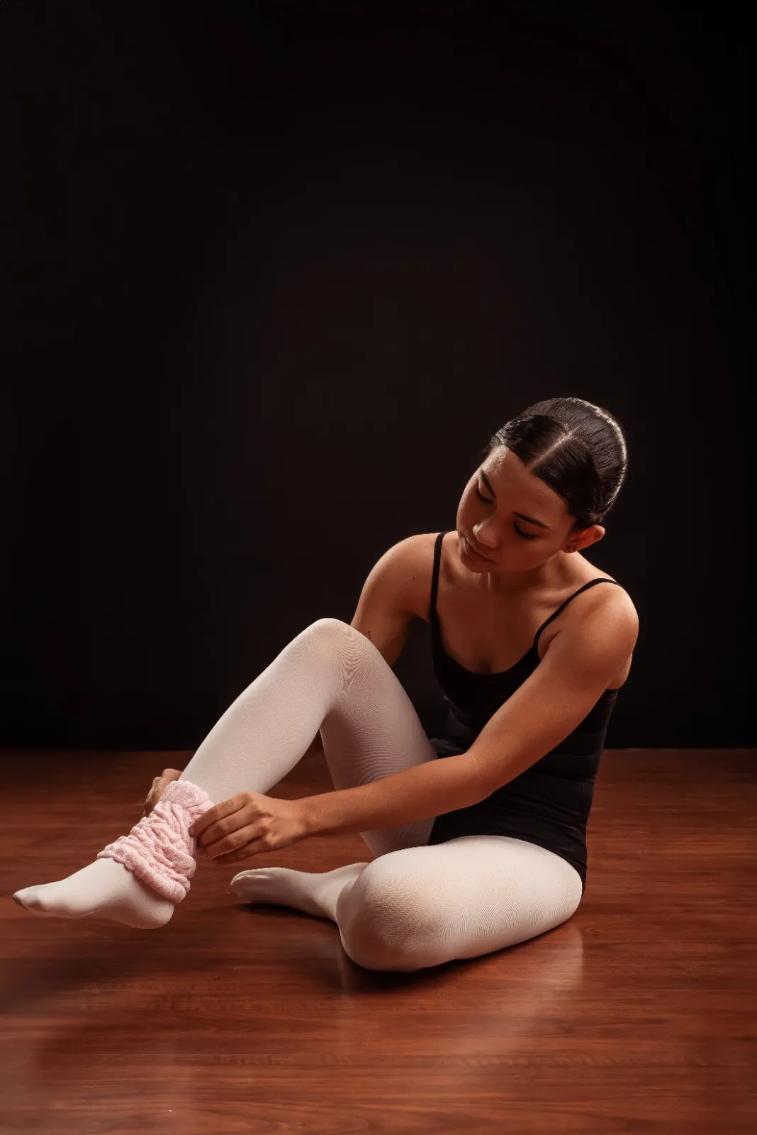 Bailarina sentada no chão ajustando a polaina rosa sobre a meia-calça branca.