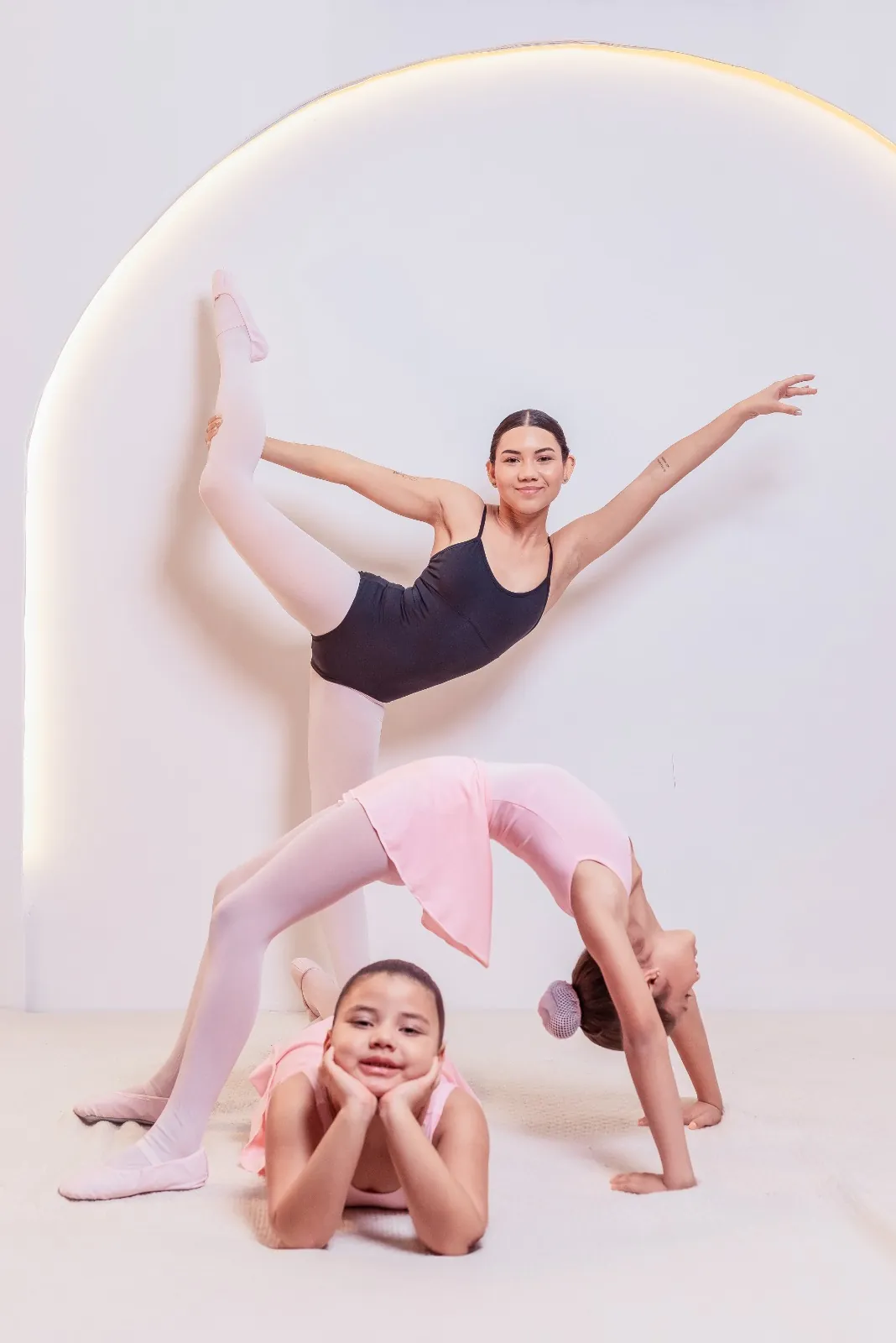 Três bailarinas da MILEVE posando em estúdio: uma jovem em arabesque, uma menina em ponte e outra deitada sorrindo, todas com collant e sapatilhas.
