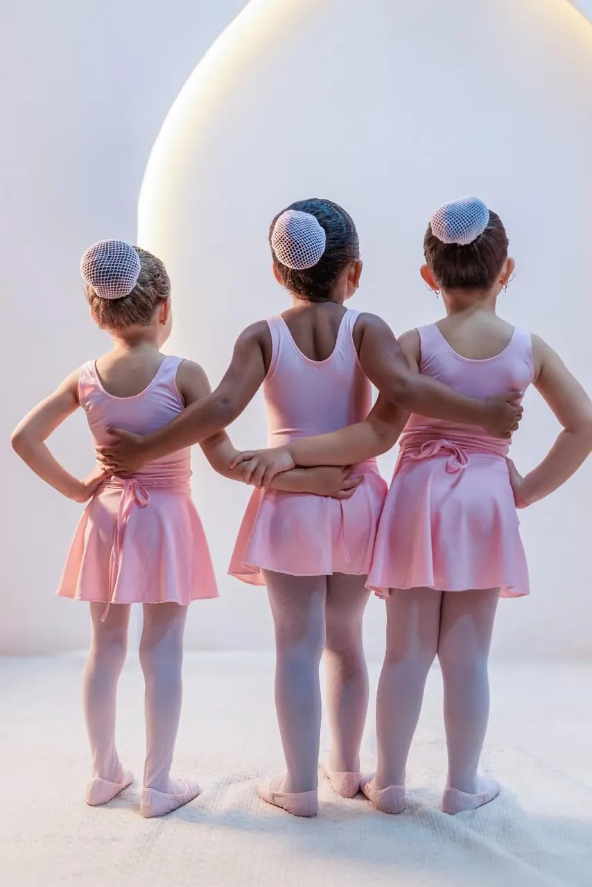 Três meninas de costas, abraçadas, usando collant rosa, saia, meia-calça e sapatilhas MILEVE.