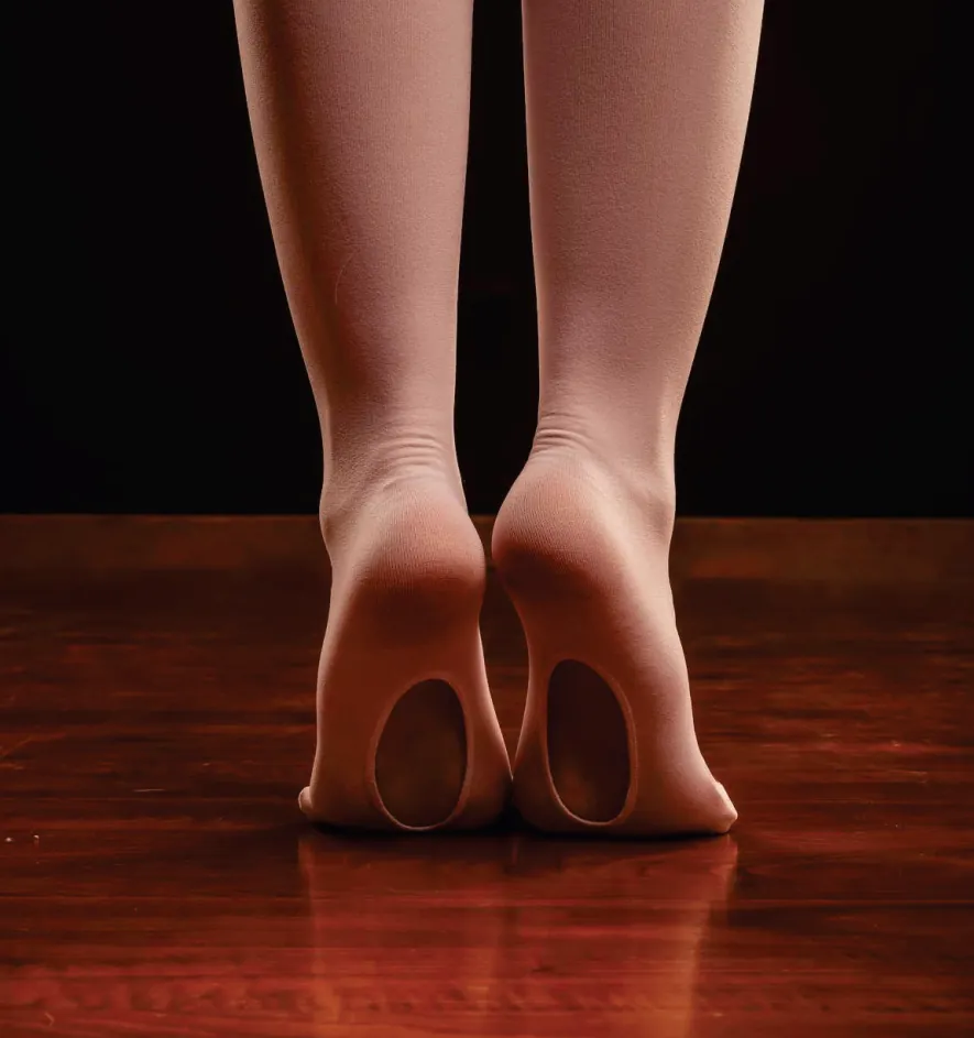 Pés de bailarina na ponta, usando meia-calça sem pé, sobre piso de madeira.